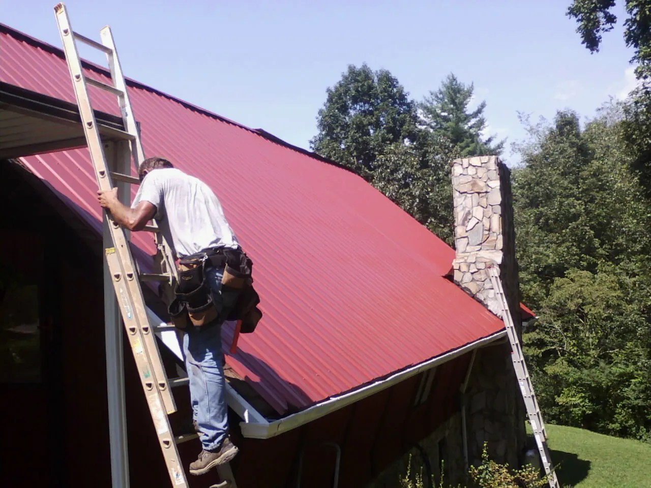 Roofer on a ladder beside a red metal roof and stone chimney — Soffit & Fascia Repair by Roofing Brothers in Poulsbo